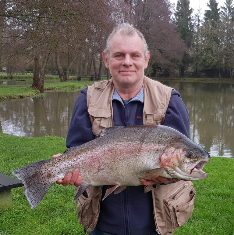 Pêcheur en gilet présentant une grosse truite arc-en-ciel devant l'étang