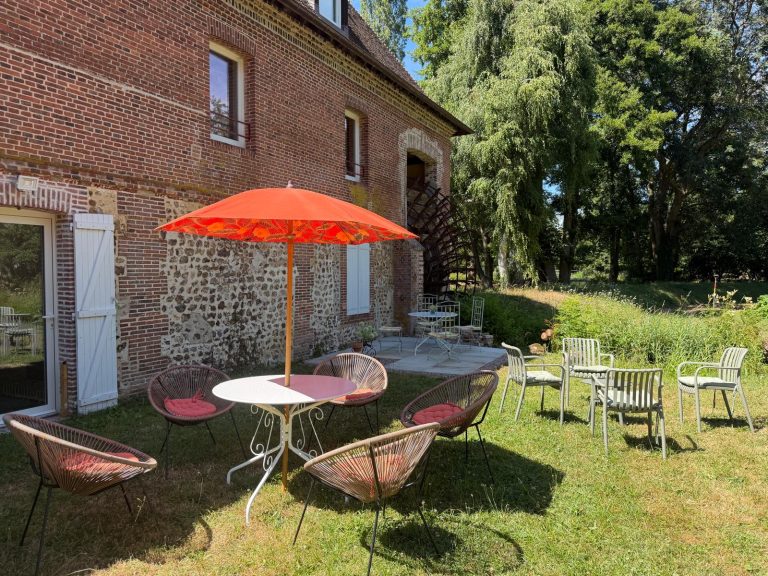 Salon de jardin et parasol orange devant la façade de brique et silex, roue du moulin à droite