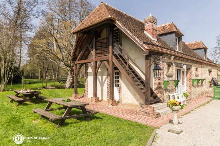 Le bâtiment normand en pierre et brique du domaine, avec son escalier extérieur couvert en charpente, ses tables de pique-nique sur la pelouse et le parc boisé en arrière-plan.