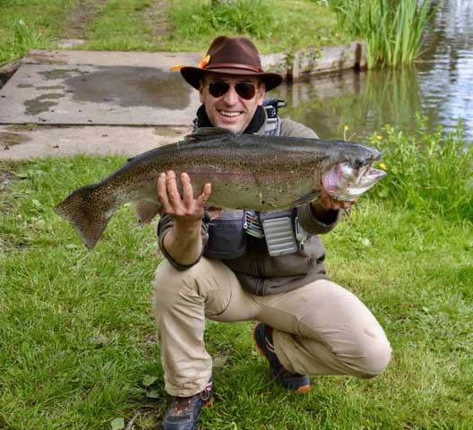 Pêcheur à la mouche agenouillé dans l'herbe, chapeau et lunettes, tenant une truite