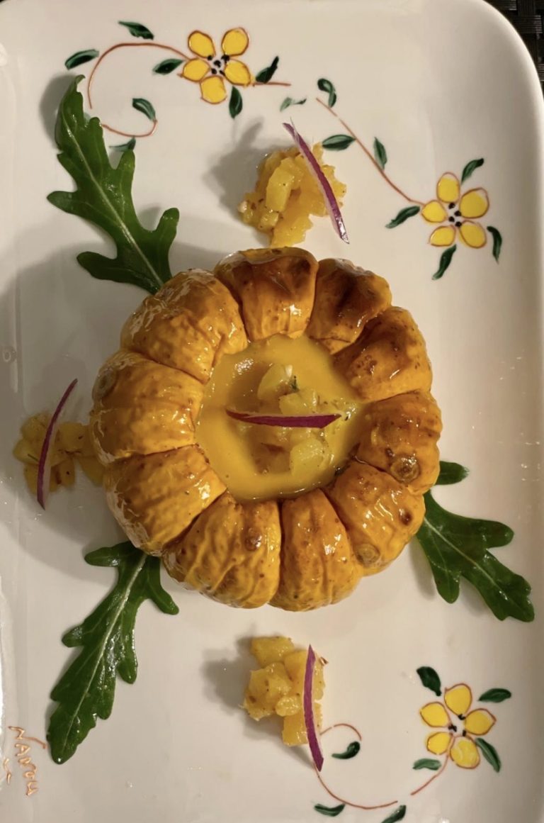 Petite courge farcie et son velouté safrané, éclats d’oignon rouge et pousses de roquette, sur une assiette à décor floral peint à la main