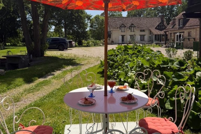 Petit-déjeuner dressé au jardin sous un parasol orange, face au corps de ferme normand